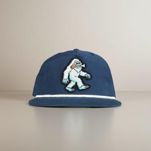Könnte beinhalten: Marineblaue Baseballkappe mit weißem Seildetail und einem Aufnäher eines Cartoon-Yetis. Der Yeti ist hellblau mit weißem Fell und schwarzen Umrissen und hält einen Knochen. Die Kappe hat einen gebogenen Schirm.