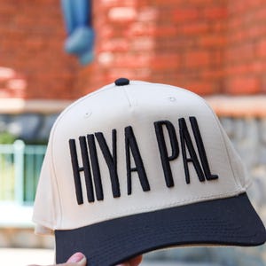 May include: A cream-coloured baseball cap with a black brim and the text "HIYA PAL" in bold, black letters. The cap has a black button on top. The background is a blurred brick building.