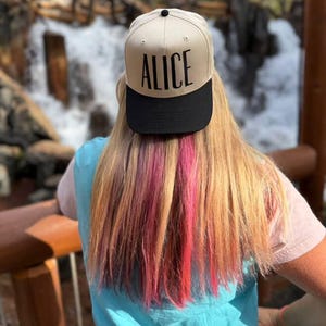 May include: A beige and black baseball cap with the name "ALICE" printed in black. The cap is worn by a person with long blonde hair with pink and red streaks. The person is wearing a blue shirt.