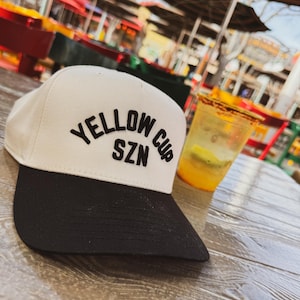May include: A white and black baseball cap with the text "Yellow Cup SZN" embroidered on the front. The cap is sitting on a wooden table.