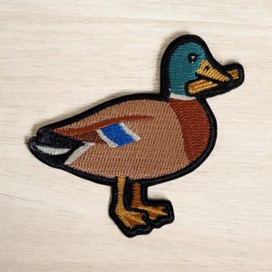 May include: Embroidered patch of a mallard duck. The duck features a teal head, brown body, and a blue wing patch. The patch is outlined in black and has yellow feet and beak.