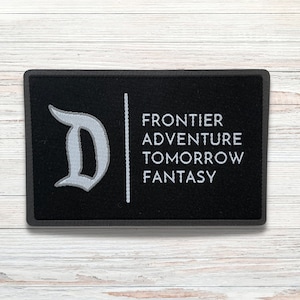 May include: Black fabric patch with a white embroidered 'D' and the words 'Frontier Adventure Tomorrow Fantasy' on a white background.