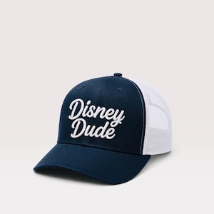 May include: A navy blue trucker hat featuring a white mesh back and a solid navy blue front panel. The text Disney Dude is embroidered in white script lettering across the front of the cap, which includes a curved brim and a button on top.