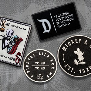 May include: Four embroidered patches featuring Disney characters and themes. The patches are black and white with white stitching. The patches include a King of Hearts playing card with Jack Skellington and Sally, a Mickey Mouse logo with the text "Mickey & Co. Est. 1928", a Pirates of the Caribbean logo with the text "A Pirates Life For Me Yo Ho Yo Ho", and a Disney logo with the text "Frontier Adventure Tomorrow Fantasy".