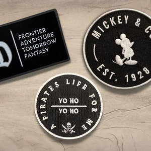 May include: Three black and white embroidered patches. The first patch is a black circle with white text that reads "A Pirates Life For Me" and a skull and crossbones. The second patch is a black circle with white text that reads "Mickey & Co. Est. 1928" and a white silhouette of Mickey Mouse. The third patch is a black rectangle with white text that reads "Frontier Adventure Tomorrow Fantasy" and a white letter "D".