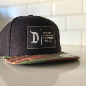 Könnte beinhalten: Schwarze und grüne Camouflage-Baseballkappe mit schwarzer Krone und einem schwarz-weißen Patch mit der Aufschrift "D" und "FRONTIER ADVENTURE TOMORROW FANTASY".