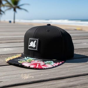 May include: A black baseball cap with a flat brim featuring a colorful tropical floral and pineapple print. The front panel displays a square black patch with a white graphic of a hand making a shaka sign gesture centered on the crown.
