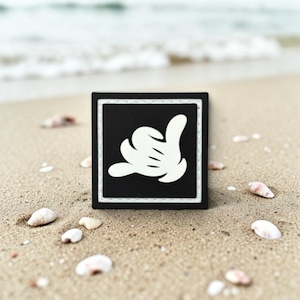 May include: A square black patch with a white border and a white graphic of a cartoon hand making a shaka gesture. The patch features a clean, bold design with a high-contrast color scheme, showcasing a stylized hand with four fingers and a thumb.