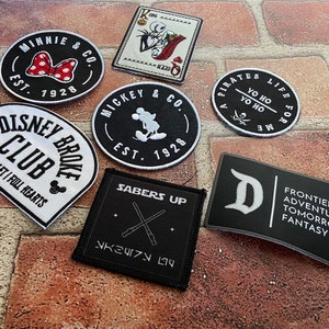 Puede incluir: Una colección de nueve parches bordados con varios personajes y temas de Disney. Los parches son negros, blancos y rojos con texto blanco. Los parches incluyen Mickey Mouse, Minnie Mouse, Piratas del Caribe, Disney Club y Star Wars.