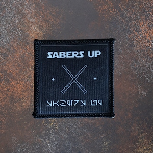 May include: Black square patch with the words "SABERS UP" in white, above crossed lightsabers. Below the lightsabers is text in a stylized font. The patch has a black border and is on a textured brown surface.