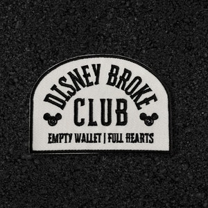 May include: White fabric patch with black embroidery. The patch reads "Disney Broke Club" with Mickey Mouse ears on either side of the text. Below the text, it reads "Empty Wallet | Full Hearts".