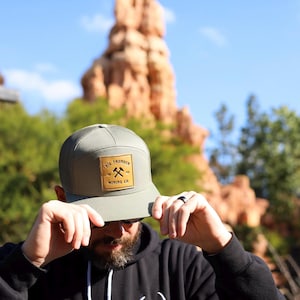 May include: Olive green flat brim cap with a brown leather patch. The patch features the text "Big Thunder Mining Co." and crossed pickaxes. The cap has a mesh back and a structured front.
