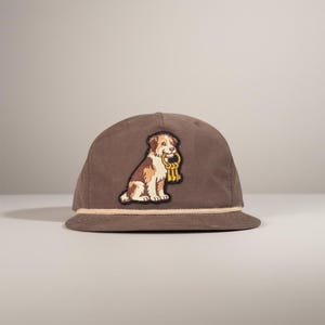 May include: A brown baseball cap with a cream-colored rope detail along the brim. The cap features an embroidered patch of a dog holding a gold key ring. The dog is brown and white.