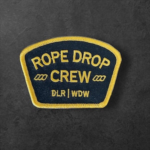 May include: A navy blue embroidered patch with a yellow border. The patch reads "ROPE DROP CREW" in yellow text, with "DLR | WDW" at the bottom. The patch has a unique shape.