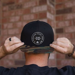 May include: A black and camouflage baseball cap with a white circle logo that says "A Pirates Life For Me" and "Yo Ho Yo Ho" inside the circle.
