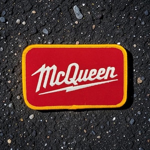 May include: A rectangular embroidered patch with a red background and a thick yellow border. The word McQueen is stitched in white cursive lettering across the center, featuring a stylized lightning bolt underline extending from the letter M.