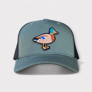 Könnte beinhalten: Eine grüne und schwarze Trucker-Kappe mit einem Entenaufnäher. Der Entenaufnäher zeigt eine Cartoon-Ente mit blauem Kopf, braunem Körper und gelbem Schnabel und Füßen. Die Kappe hat einen gebogenen Schirm und eine Mesh-Rückseite.