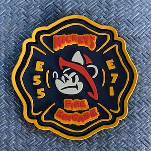 May include: A navy blue and yellow fire department badge with the text "Mickey's Fire Brigade". The badge features a cartoon character with a red hat and a fire axe. The badge also has the numbers "E55" and "E71".