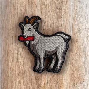 May include: Embroidered patch featuring a gray goat with a red object in its mouth. The goat has brown horns and a black outline. The patch is on a wooden surface.