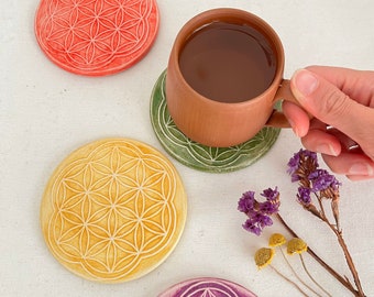 May include: Four ceramic coasters with a flower of life design in different colors: red, yellow, green, and purple. A brown ceramic mug with a dark brown liquid inside is sitting on the green coaster.