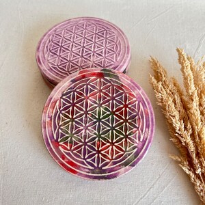May include: Two round, purple coasters with a white, geometric flower of life design. The bottom coaster has red, green, and purple paint. The coasters are made of a textured material.