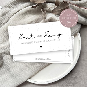 May include: A white card with black script that reads "Zeit statt Zeug" and "Das sch&ouml;nste Geschenk ist gemeinsame Zeit." A small heart is below the text. The card is on a white plate and a gray cloth.