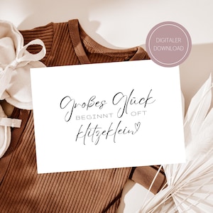 Puede incluir: Un body de bebé acanalado marrón con una tarjeta blanca con el texto "Großes Glück beginnt oft Hitzeklein" en escritura negra. Se ve una etiqueta de descarga digital. La imagen tiene una paleta de colores suaves y neutros.