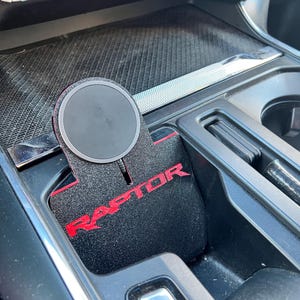 Puede incluir: Una plataforma de carga inalámbrica negra con un logotipo "RAPTOR" rojo en una alfombra interior de coche negra.