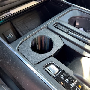 May include: A close-up of a black car's centre console with a cup holder, gear shift, and a button labelled "000".