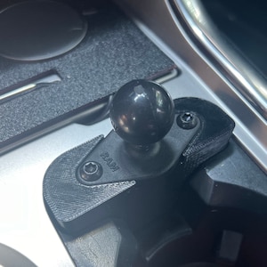 以下が含まれることがあります： 車のセンターコンソールに取り付けられた、黒いRAMボールマウントと黒いボールヘッド。マウントには白い文字で「RAM」と表示されています。