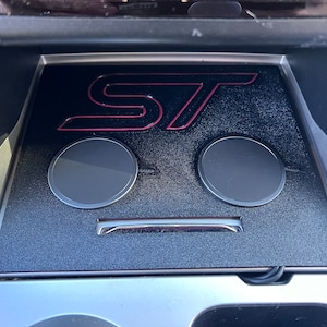 Puede incluir: Un interior de coche negro con una plataforma de carga inalámbrica que presenta el logotipo "ST" en rojo. La plataforma de carga tiene dos áreas de carga circulares.