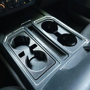 May include: A black plastic cup holder insert for a car. The insert has two cup holders on the left and two on the right. The insert is designed to fit into the centre console of a car.