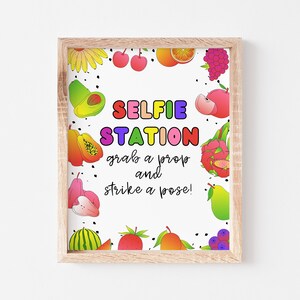 May include: A framed printable sign with a white background and a colorful fruit border. The sign says "SELFIE STATION grab a prop and strike a pose!"