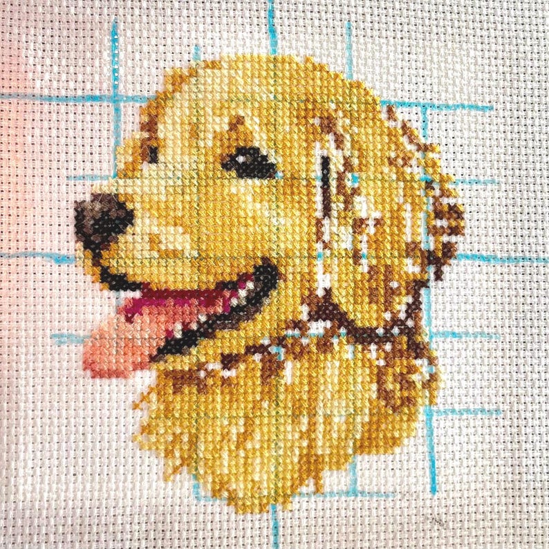 K&ouml;nnte beinhalten: Kreuzstich-Stickerei eines Golden Retriever Hundes mit herausgestreckter rosa Zunge. Der Hund ist auf einem wei&szlig;en Aida-Stoff mit einem blauen Gittermuster gestickt.