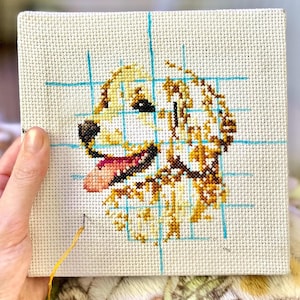 K&ouml;nnte beinhalten: Eine Nahaufnahme einer Kreuzstich-Stickerei eines Golden Retriever-Hunde-Kopfes. Der Hund l&auml;chelt mit herausgestreckter Zunge. Die Stickerei befindet sich auf einem wei&szlig;en Stoff mit blauen Gitterlinien.