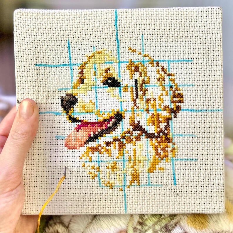 K&ouml;nnte beinhalten: Eine Nahaufnahme einer Kreuzstich-Stickerei eines Golden Retriever-Hunde-Kopfes. Der Hund l&auml;chelt mit herausgestreckter Zunge. Die Stickerei befindet sich auf einem wei&szlig;en Stoff mit blauen Gitterlinien.