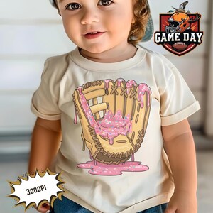 May include: A toddler wearing a light beige t-shirt with a donut-themed baseball glove graphic. The graphic is pink, brown, and white. The text "GAME DAY" is in a red and white speech bubble in the top right corner of the image.