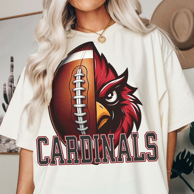 May include: Cream-coloured t-shirt with a graphic design. The design is split vertically, with a football on the left and a red cardinal bird on the right. The word "CARDINALS" is printed in a bold, block font below the graphic.