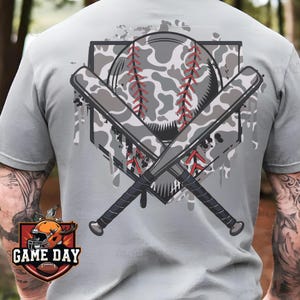 May include: A gray t-shirt with a camouflage baseball and crossed bats design. The design has a white and gray camouflage pattern with red stitching. The text "GAME DAY" is on a red and orange badge in the lower left corner.