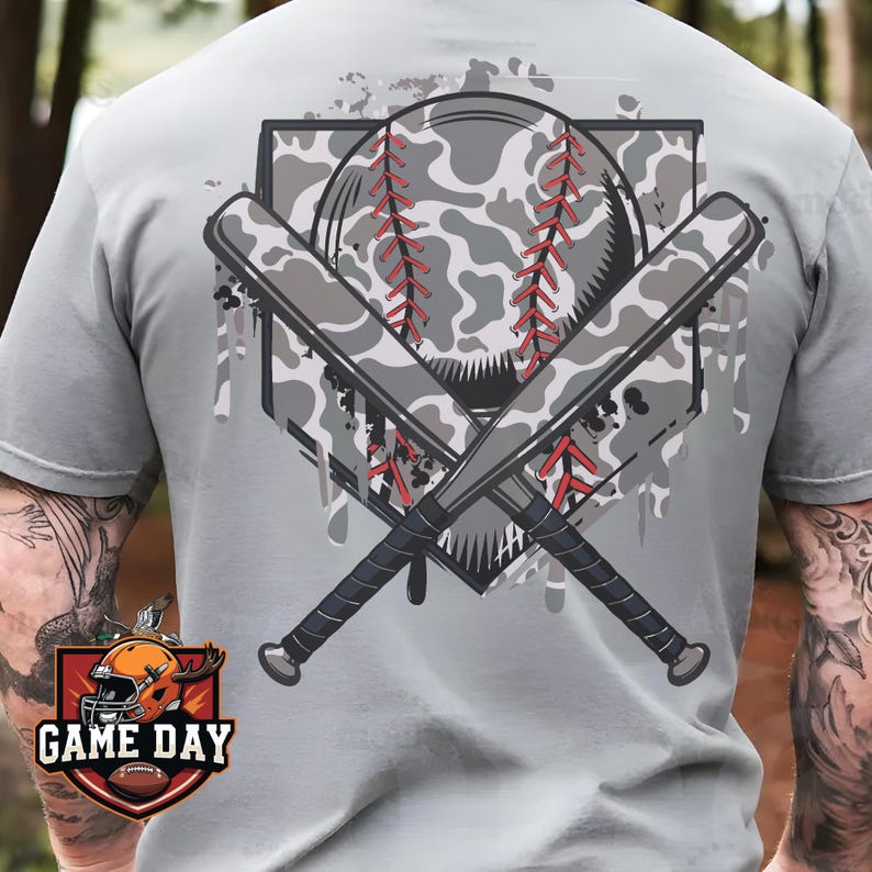 May include: A gray t-shirt with a camouflage baseball and crossed bats design. The design has a white and gray camouflage pattern with red stitching. The text "GAME DAY" is on a red and orange badge in the lower left corner.
