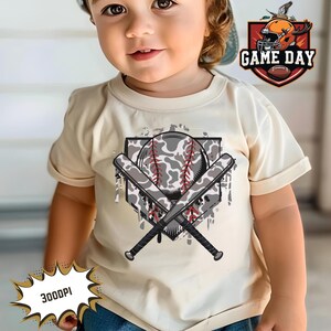 May include: A toddler wearing a cream-colored t-shirt with a baseball graphic. The graphic features a camouflage baseball with two crossed bats and a red and white baseball diamond. The text "GAME DAY" is in a red and white banner above the graphic.