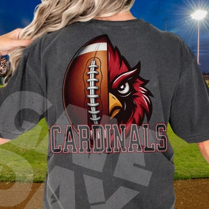 May include: Charcoal grey t-shirt with a graphic of a football and a cardinal. The football is brown with white stitching, and the cardinal is red and black. The word "CARDINALS" is printed in a bold, outlined font.