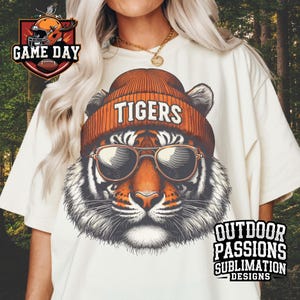 May include: A white t-shirt featuring a graphic of a tiger wearing a beanie with the word "TIGERS" on it. The tiger is wearing sunglasses. The text "OUTDOOR PASSIONS SUBLIMATION DESIGNS" is in the bottom right corner of the shirt.