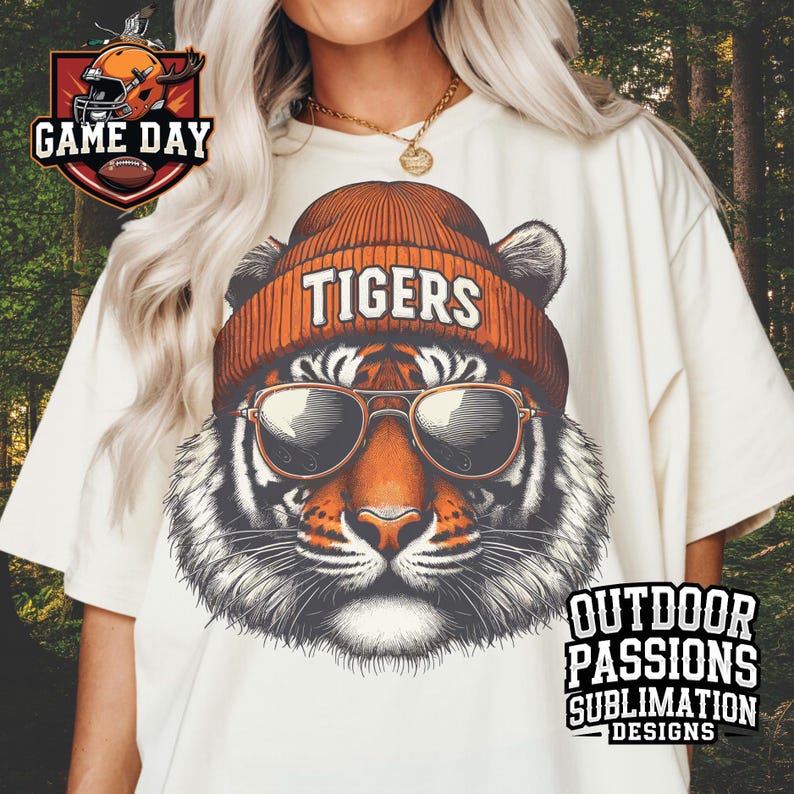 May include: A white t-shirt featuring a graphic of a tiger wearing a beanie with the word "TIGERS" on it. The tiger is wearing sunglasses. The text "OUTDOOR PASSIONS SUBLIMATION DESIGNS" is in the bottom right corner of the shirt.