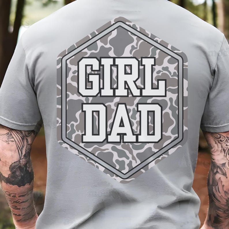 K&ouml;nnte beinhalten: Ein graues T-Shirt mit einem sechseckigen Patch auf der R&uuml;ckseite. Der Patch zeigt ein Tarnmuster in Graut&ouml;nen und Wei&szlig;. Der Text "GIRL DAD" ist in wei&szlig;en Buchstaben auf dem Patch gedruckt.