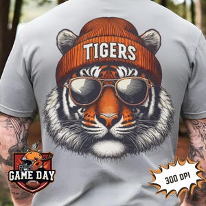 May include: A grey t-shirt featuring a graphic of a tiger wearing sunglasses and a beanie with the word "TIGERS" on it. The graphic is in orange, black, and white. The t-shirt has the words "GAME DAY" in a red and orange logo on the bottom left and "300 DPI" in a red and white logo on the bottom right.