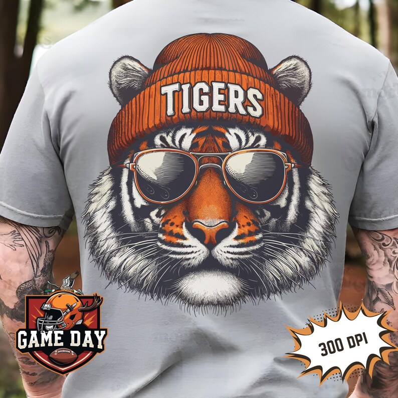 May include: A grey t-shirt featuring a graphic of a tiger wearing sunglasses and a beanie with the word "TIGERS" on it. The graphic is in orange, black, and white. The t-shirt has the words "GAME DAY" in a red and orange logo on the bottom left and "300 DPI" in a red and white logo on the bottom right.