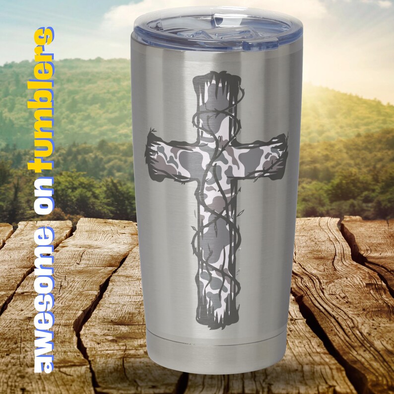 May include: A stainless steel tumbler with a camouflage cross design. The cross is surrounded by a vine-like pattern. The tumbler is sitting on a wooden surface.