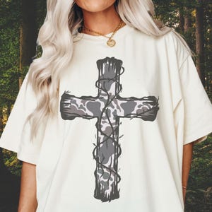 May include: A white t-shirt with a gray camouflage cross design. The cross is surrounded by a vine-like pattern.