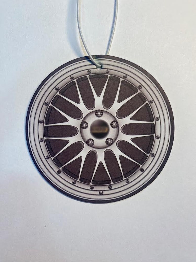May include: A circular car air freshener featuring a detailed illustration of a silver and dark gray alloy wheel. The wheel design includes multiple spokes and a central hub, suspended by a thin, light-colored string.